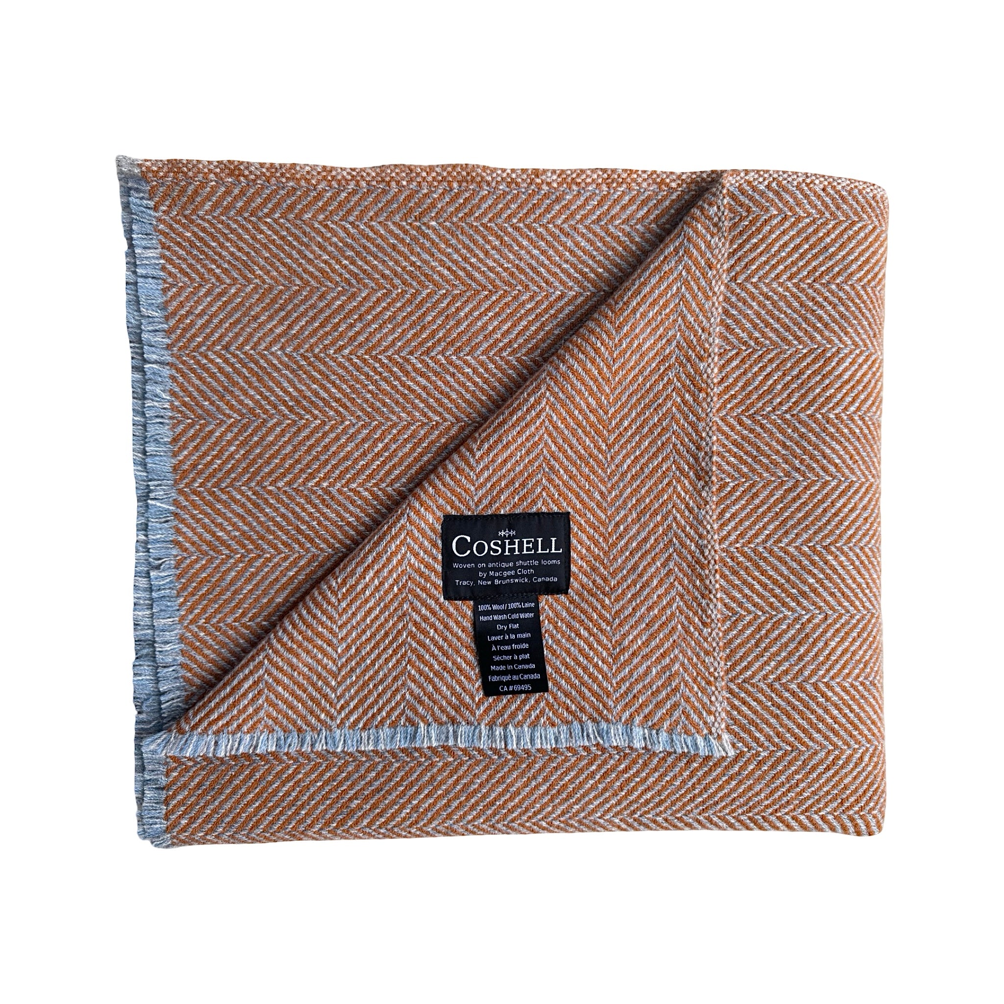 A folded herringbone pattern wool blanket by Coshell, featuring a warm brown and beige weave with a light blue-gray fringed edge and a black brand label.