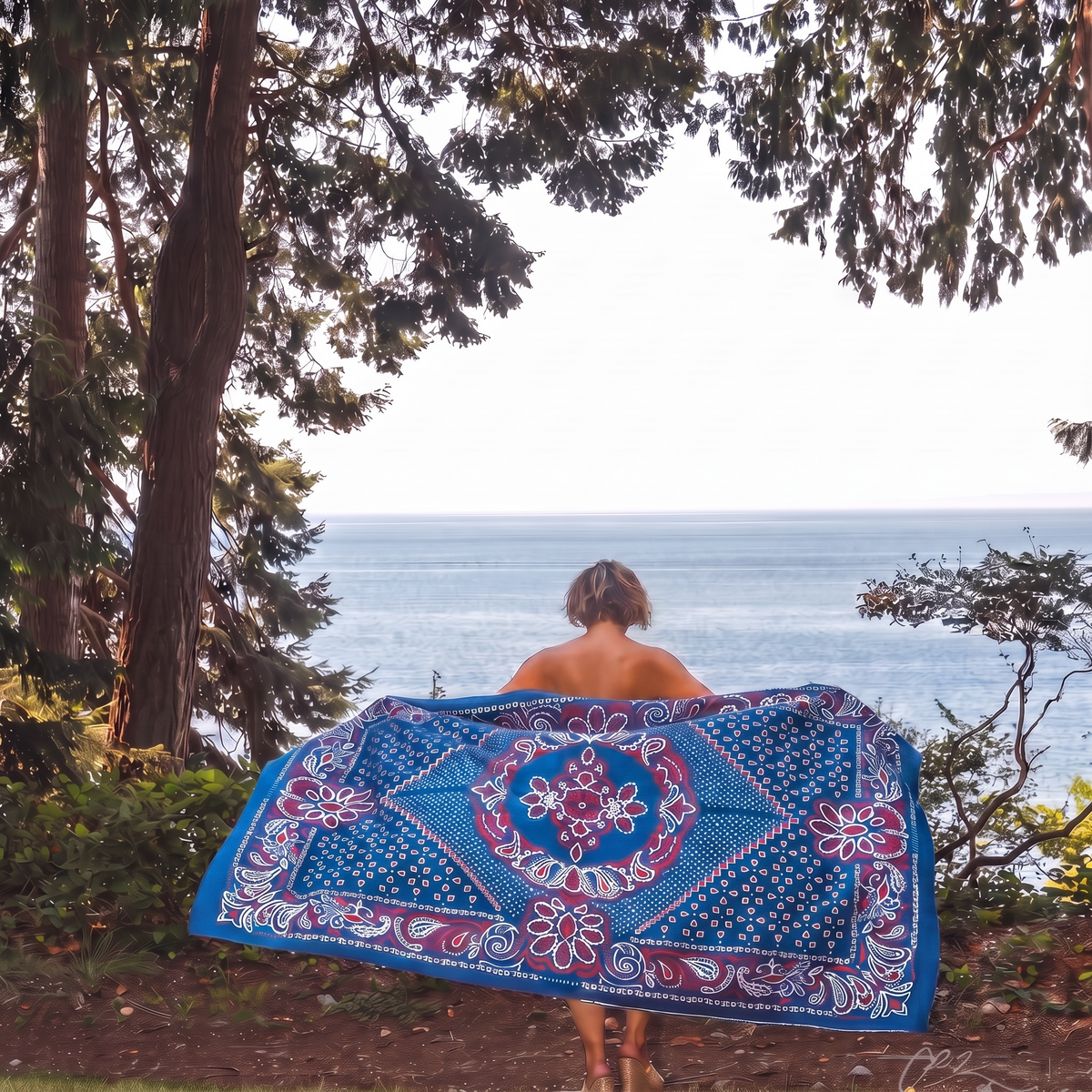 A person stands with their back to the camera holding up a large blue bandana with a red and white paisley pattern over their shoulders, with a forest and ocean in the background.