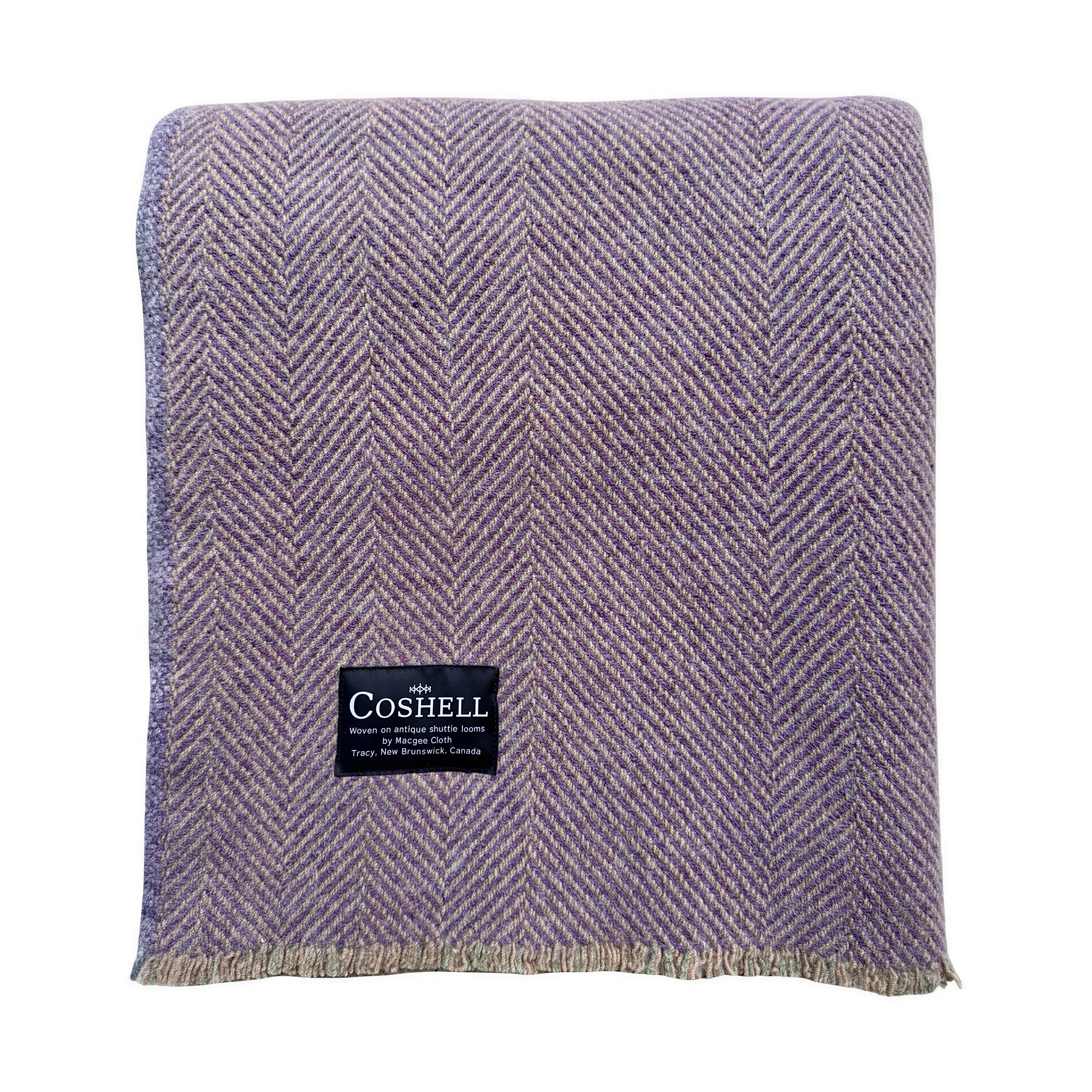 A woven throw blanket in a dusty lavender-purple orange and green weft herringbone pattern, featuring a black Coshell brand label on the front and fringed edges.
