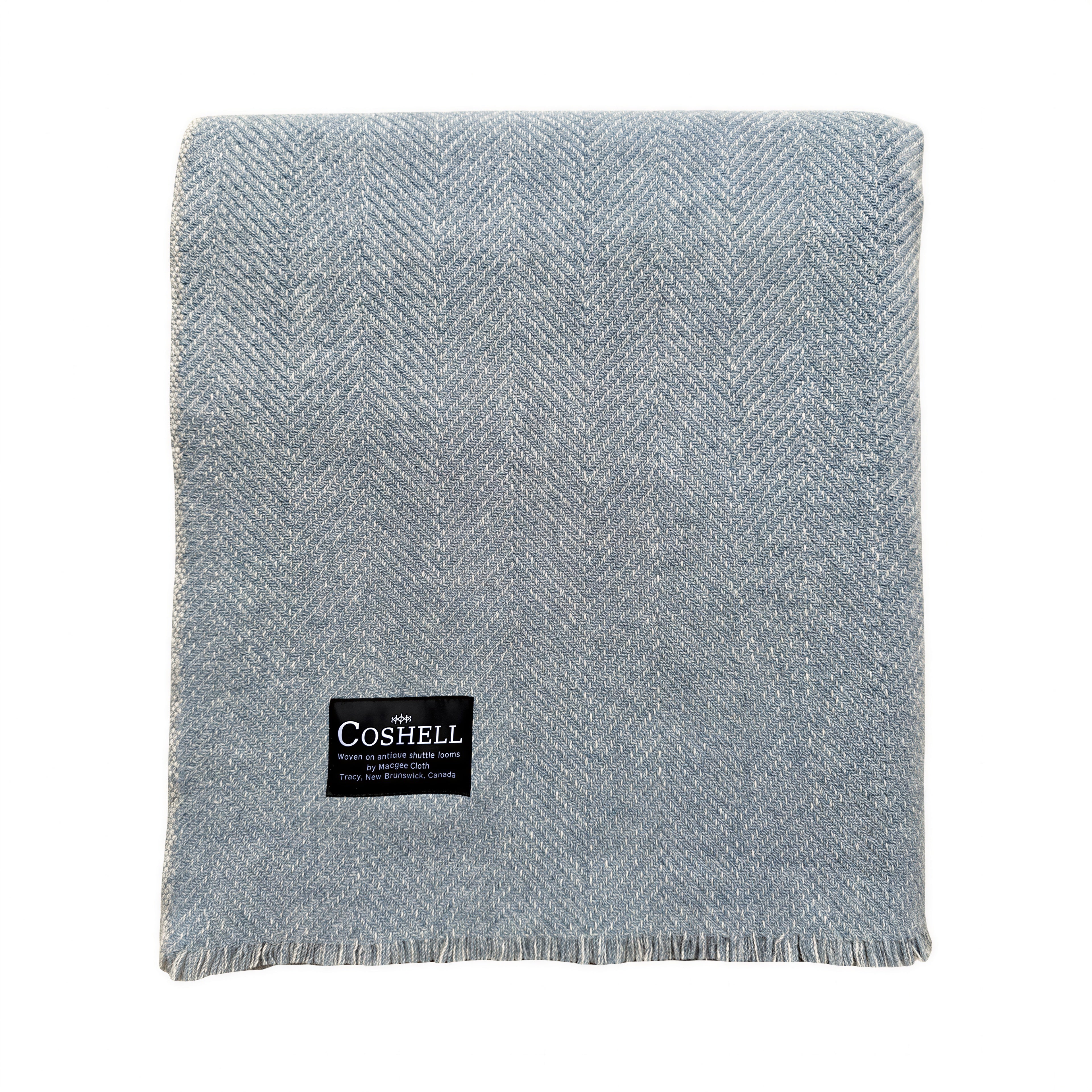 A folded light blue and grey herringbone patterned throw blanket with a black Coshell brand label on the front