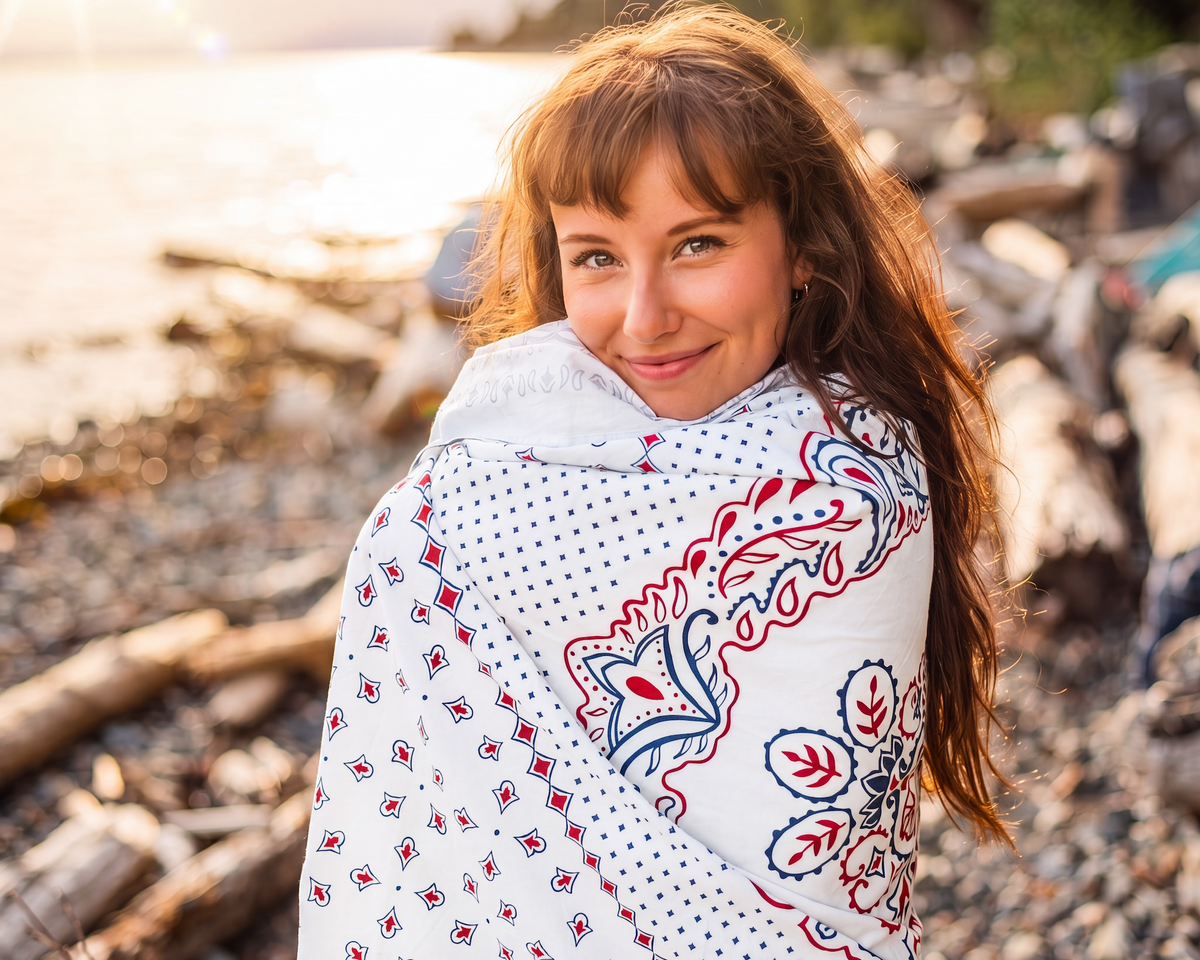 A woman with long light brown hair stands on a rocky beach at sunset, wrapped in a white hooded blanket featuring red and blue floral and geometric patterns.