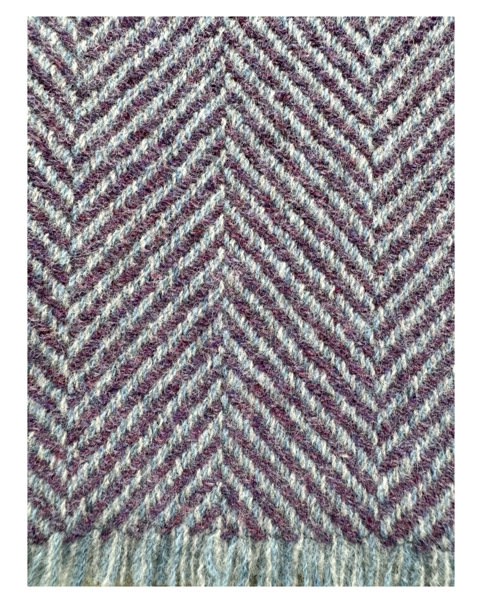 A close-up of woven wool textile with a herringbone pattern in muted purple, beige, and light blue-gray tones, with a fringed edge visible along the bottom.