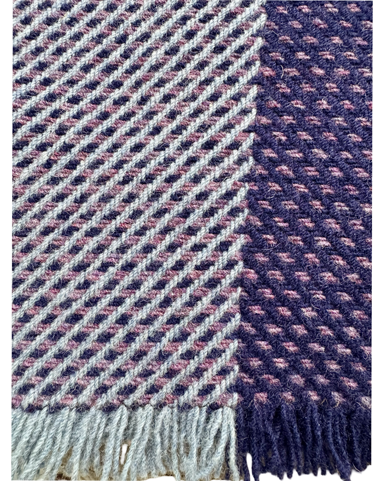A close-up of  woven wool textile with fringe, showing a diagonal herringbone pattern in light gray, navy, and muted pink, with a solid navy section on the right.