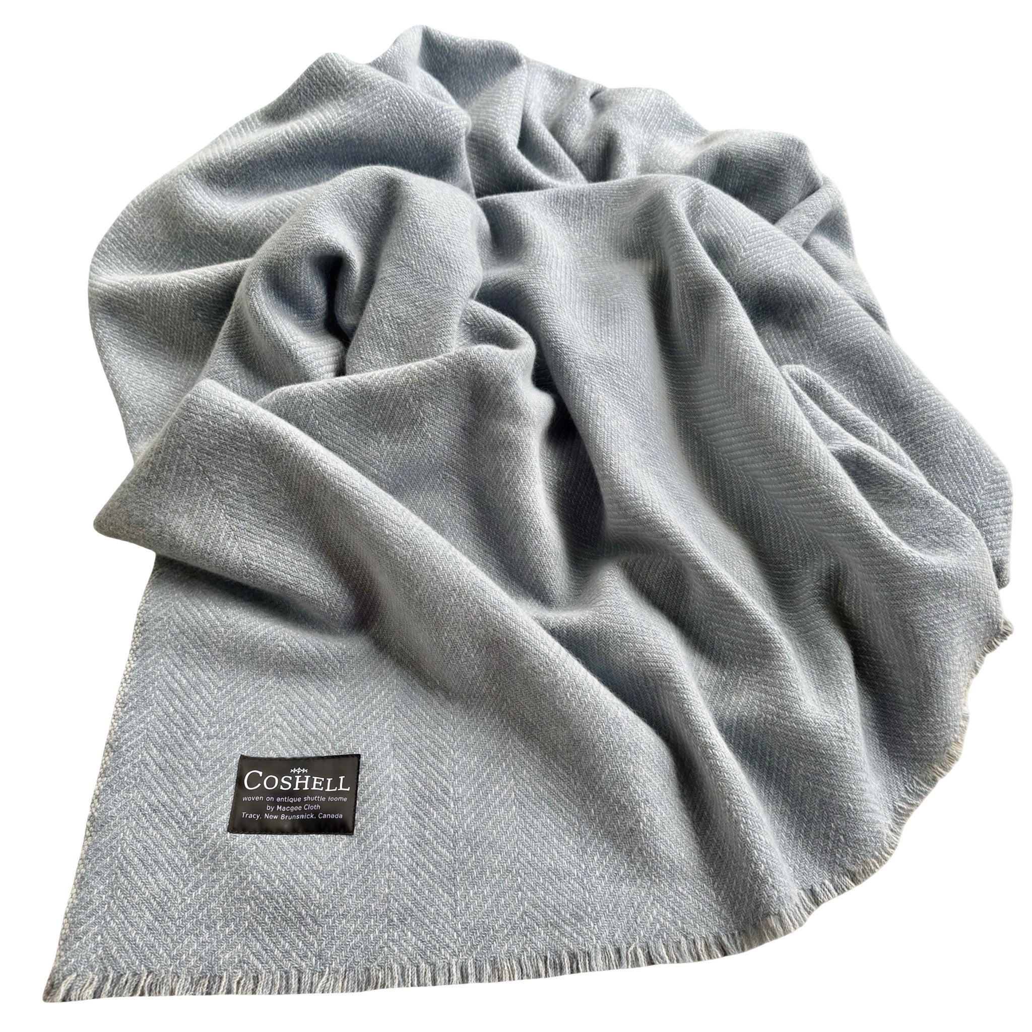 A light blue gray herringbone patterned throw blanket with fringed edges, draped to show its texture and a small black Coshell brand label near the bottom left corner