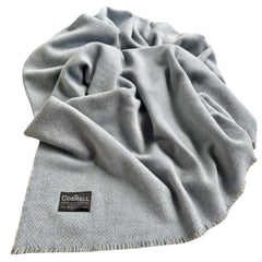 A light blue gray herringbone patterned throw blanket with fringed edges, draped to show its texture and a small black Coshell brand label near the bottom left corner
