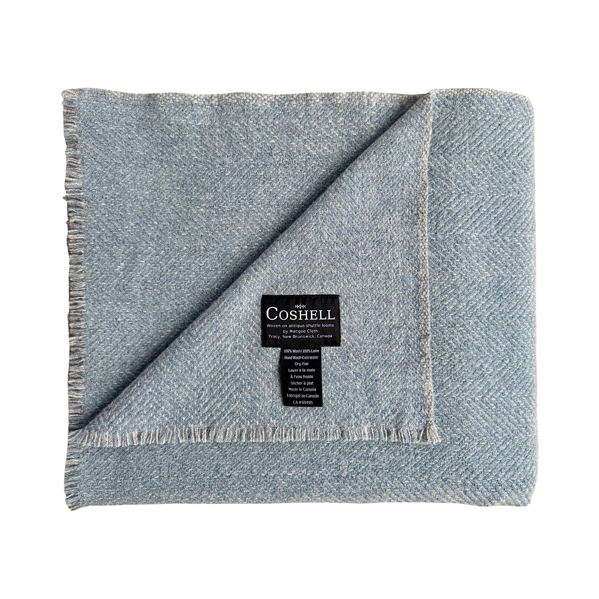 A folded light blue herringbone woven throw blanket with fringed edges, featuring a black Coshell brand label in the center, 