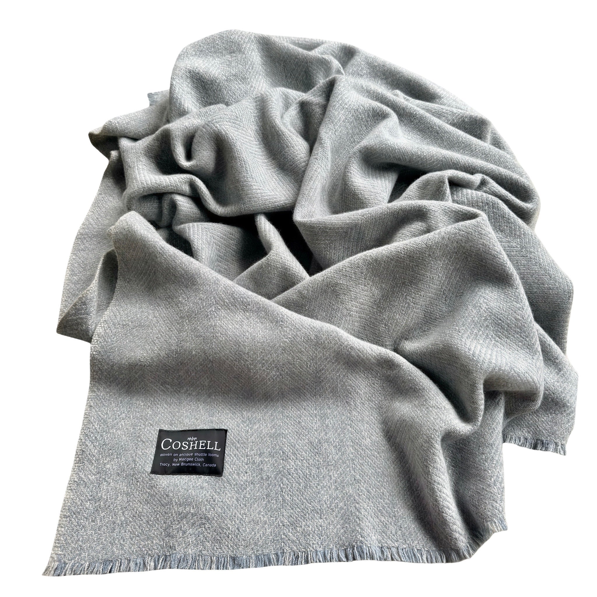 A light gray and blue woven blanket with fringed edges, draped in soft folds on a white background. A black woven brand label reading 'COSHELL' is visible near the lower left edge of the scarf.