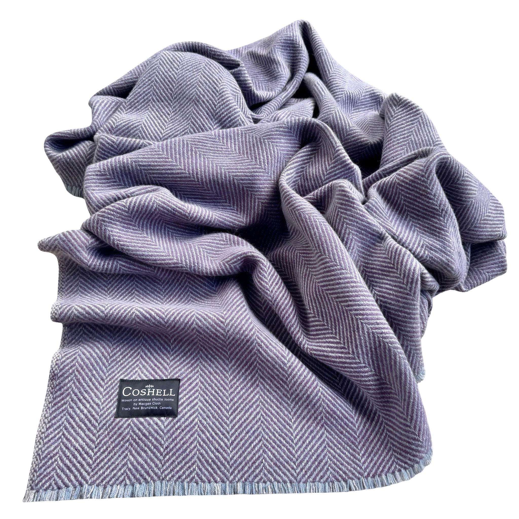 A draped herringbone pattern throw blanket in a dusty lavender-gray color, featuring a black Coshell brand label and fringed edges