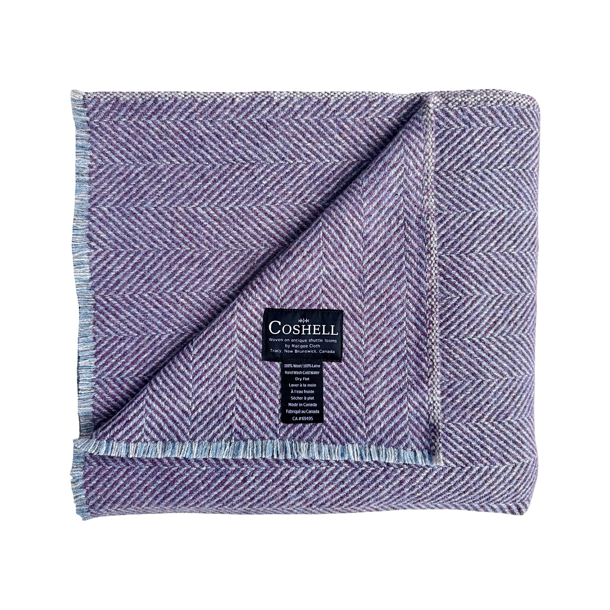 A folded herringbone pattern throw blanket in lavender purple grey and blue, featuring fringed edges and a black Coshell brand label in the center
