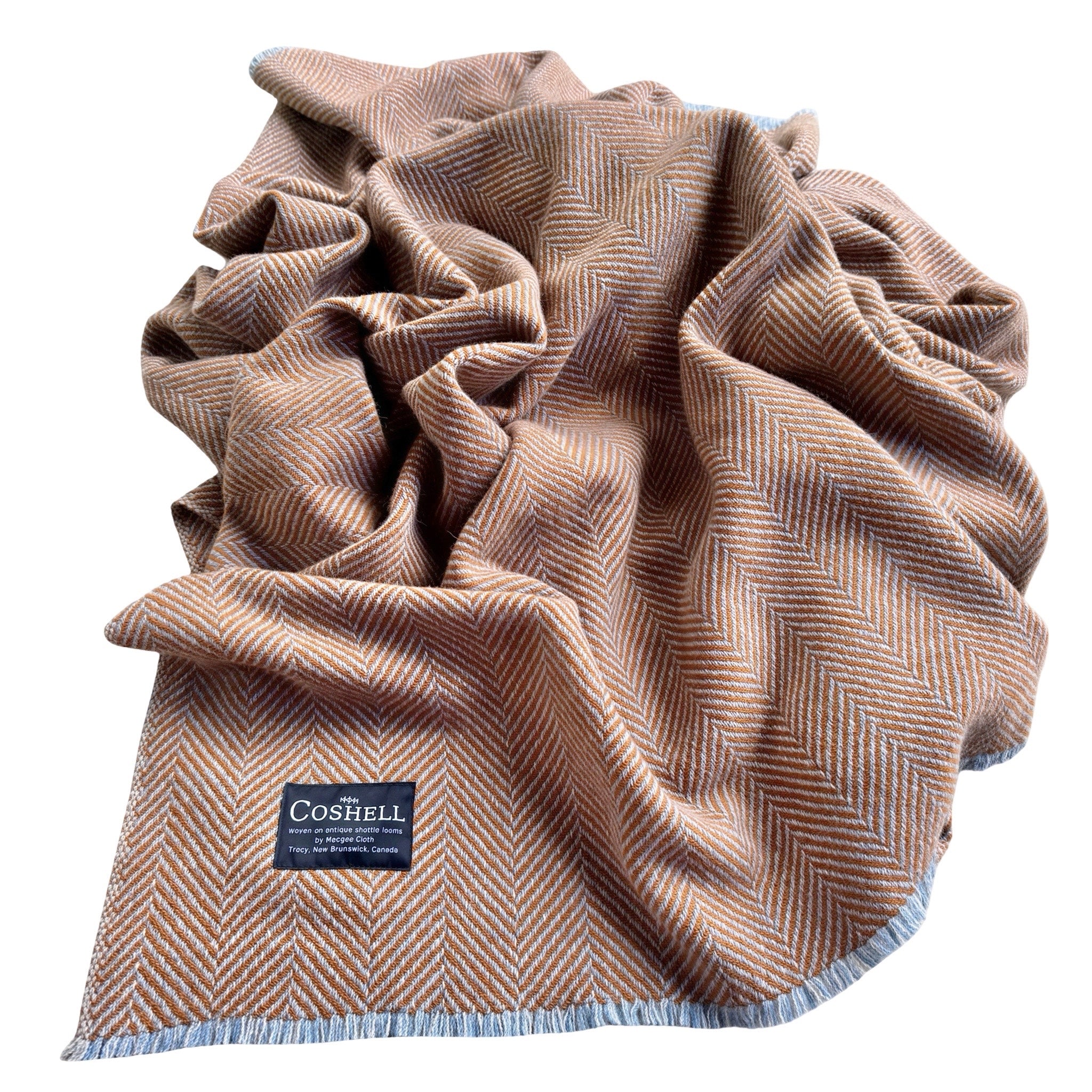 A folded herringbone pattern throw blanket in orange with light blueand grey warp fringed edges, featuring a black Coshell brand label.