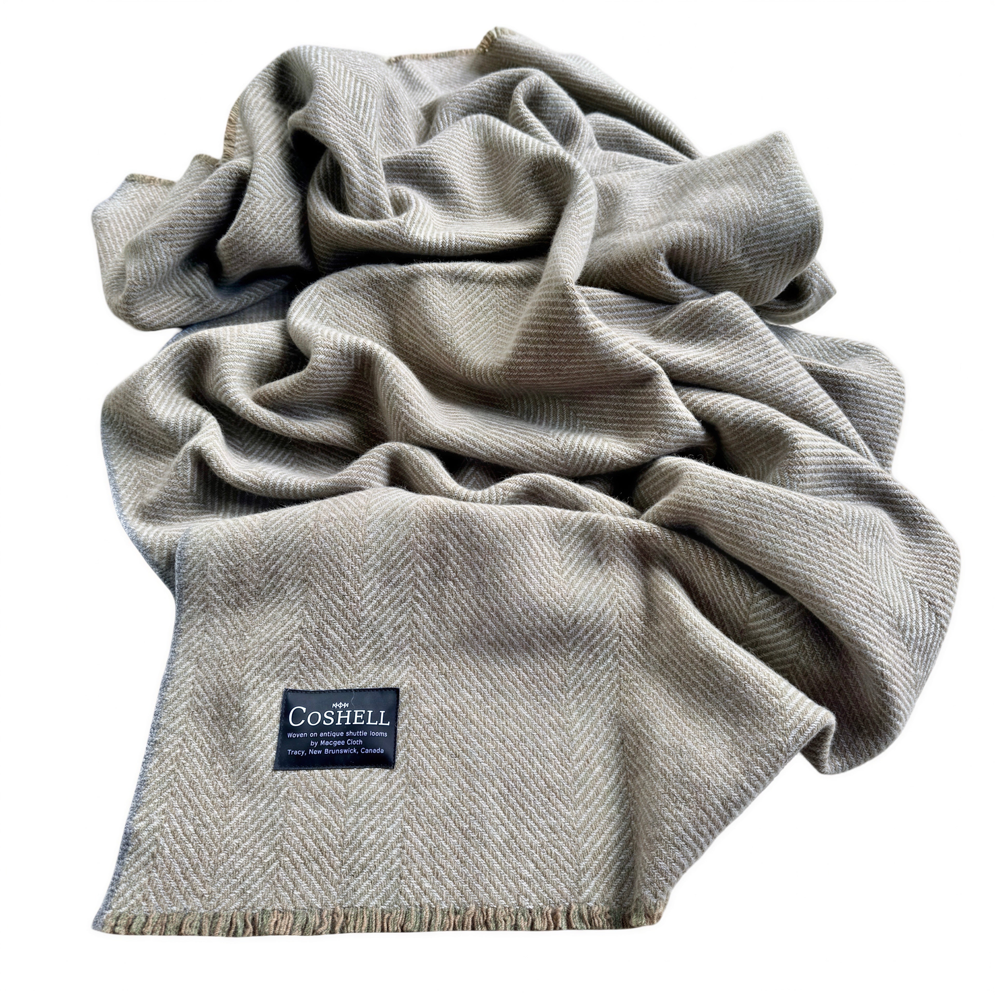 A light brown and green warp and gray herringbone patterned throw blanket with fringed edges, draped in soft folds. A black Coshell brand label is visible on the lower left corner.