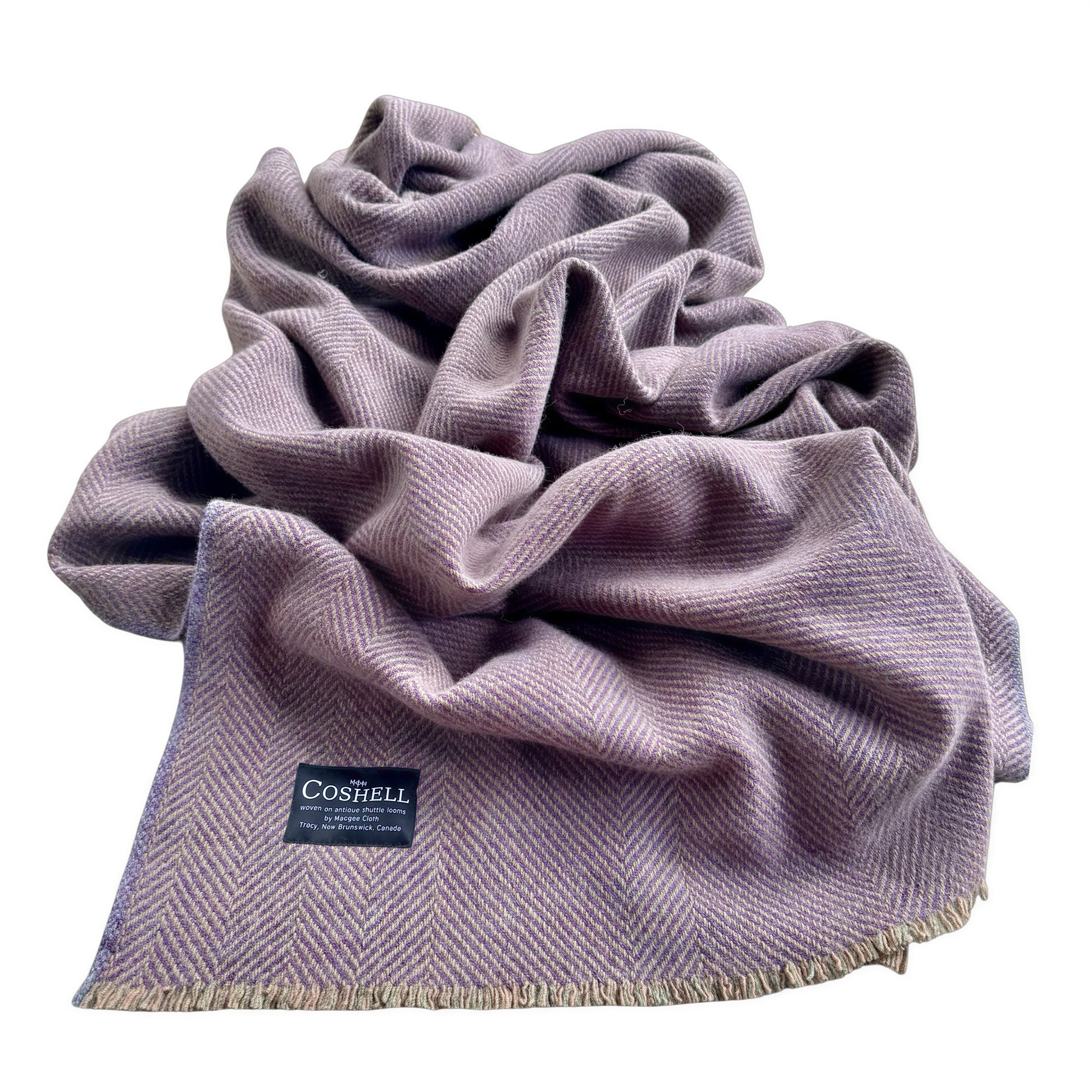 A dusty purple herringbone woven blanket with fringed edges and a black Coshell brand label