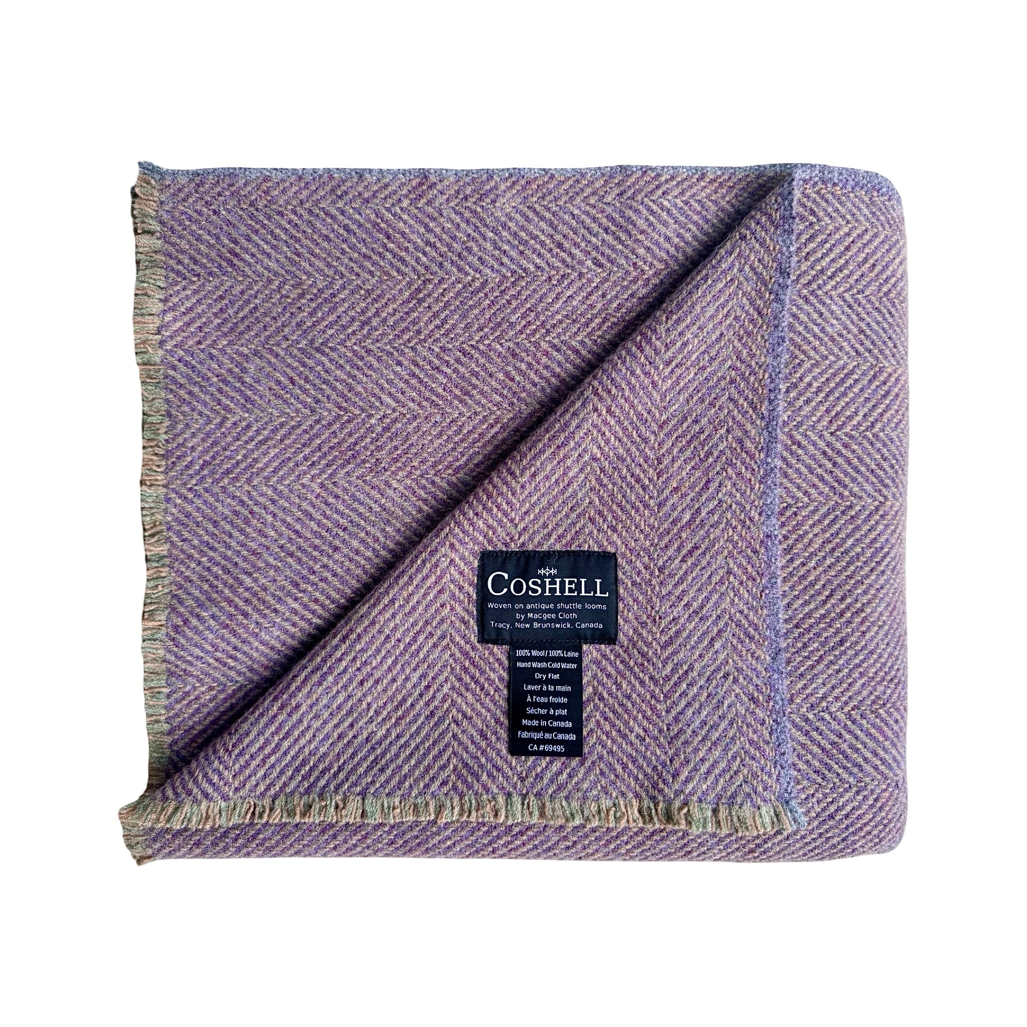 A folded herringbone patterned throw blanket in a dusty purple green and orange colorway, featuring a black Coshell brand label and fringed edges