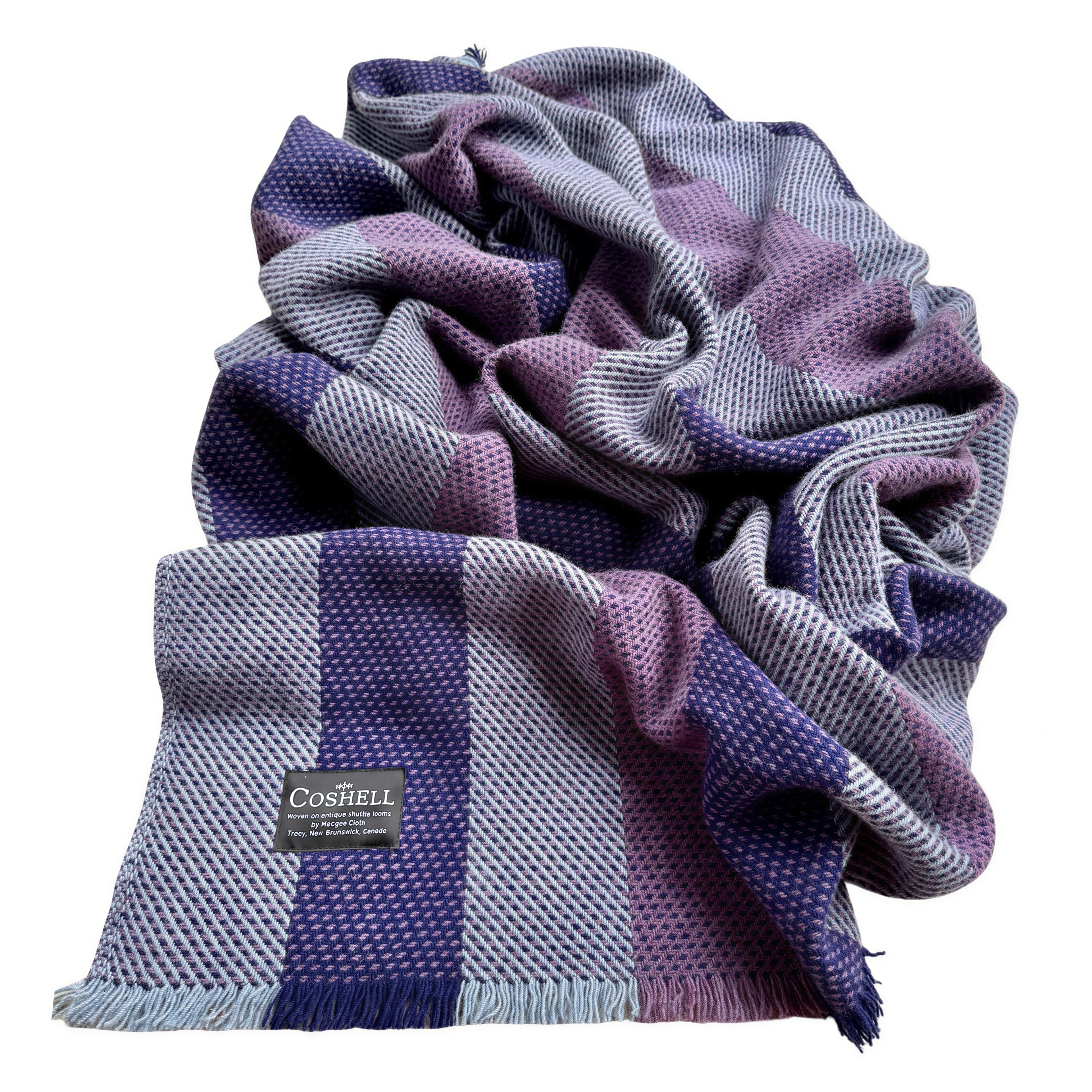 A folded and draped woven blanket from Coshell, featuring a textured checkered pattern in shades of purple, lavender, slate blue, and gray, with a small black brand label visible.