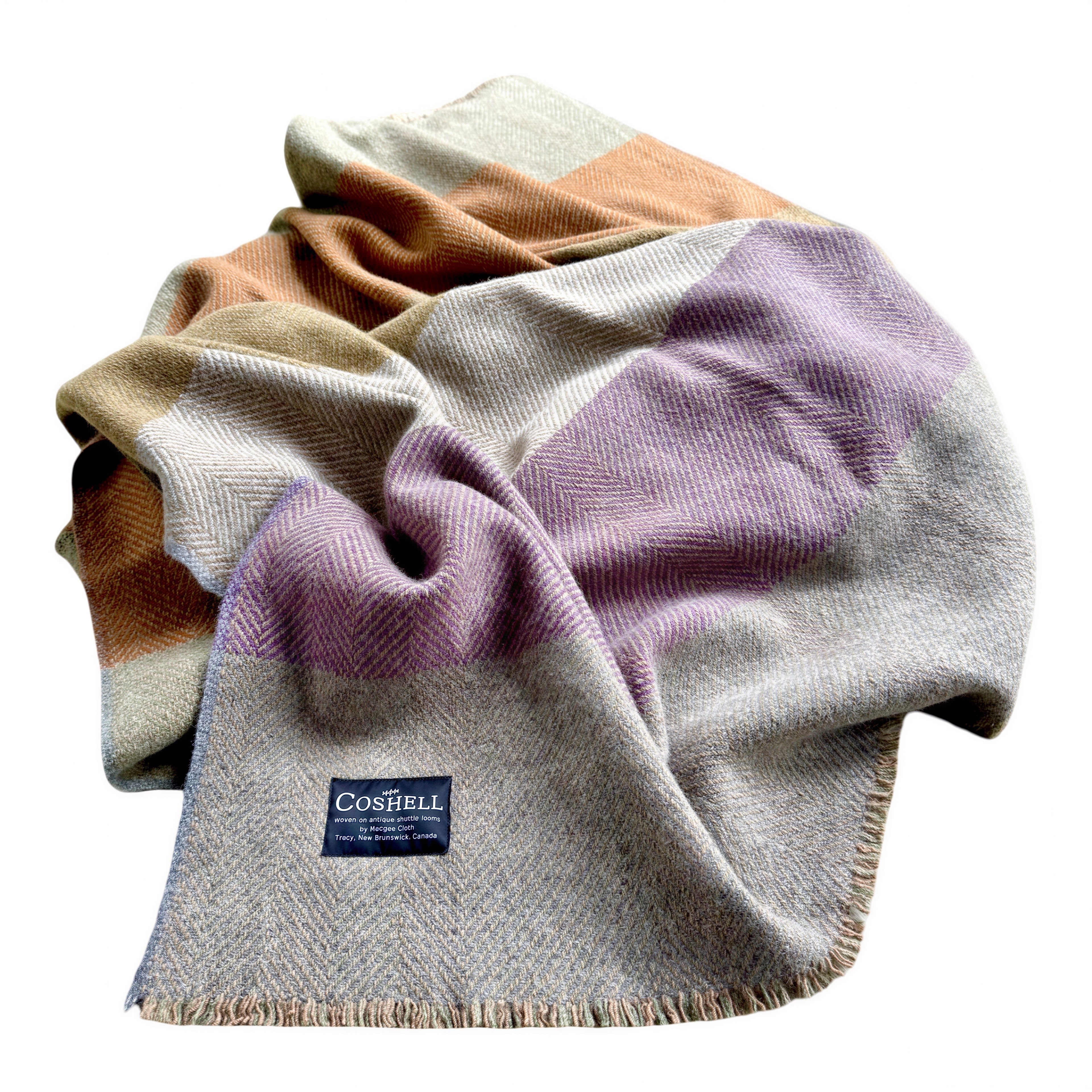 A folded herringbone wool blanket by The Coshell, featuring a color-blocked pattern in beige, dusty purple, tuscan orange, and light gray with fringed edges and a visible brand label.